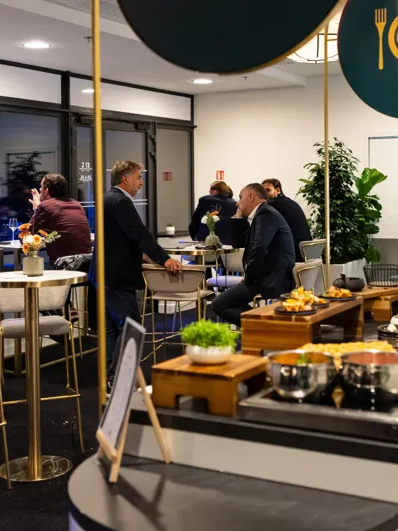 Guests in the Tie-Break lounge chatting and enjoying their drinks, with the match visible through the large window overlooking the court.
