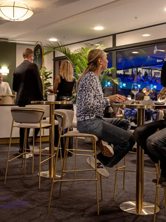 Invités du Salon Tie-Break discutant autour de leurs consommations, avec le match visible en arrière-plan via la baie vitrée.